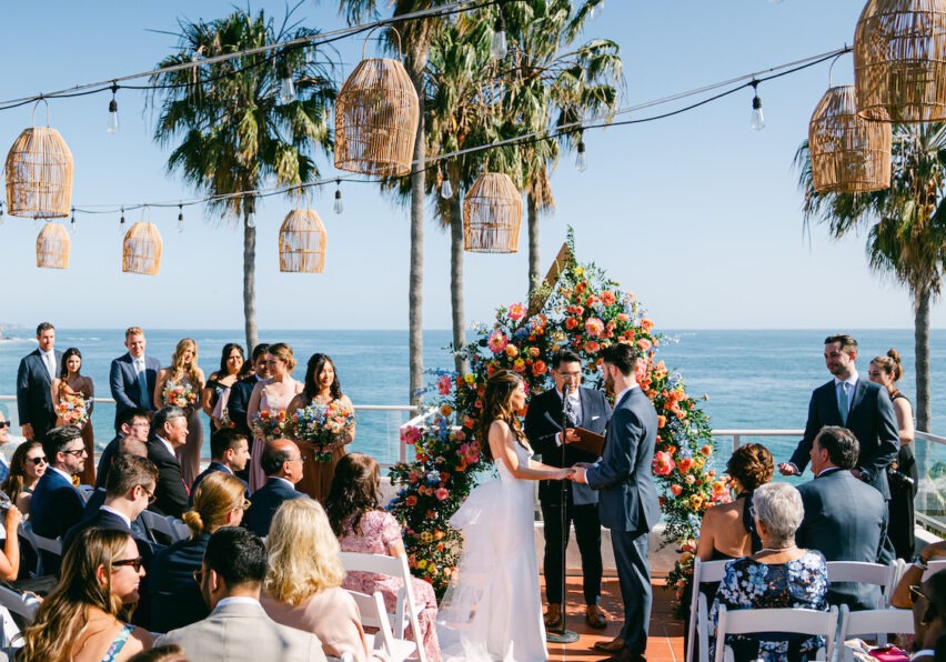 Outdoor wedding ceremony by the ocean, featuring a couple exchanging vows under a floral arch, surrounded by guests, palm trees, and decorative lights.