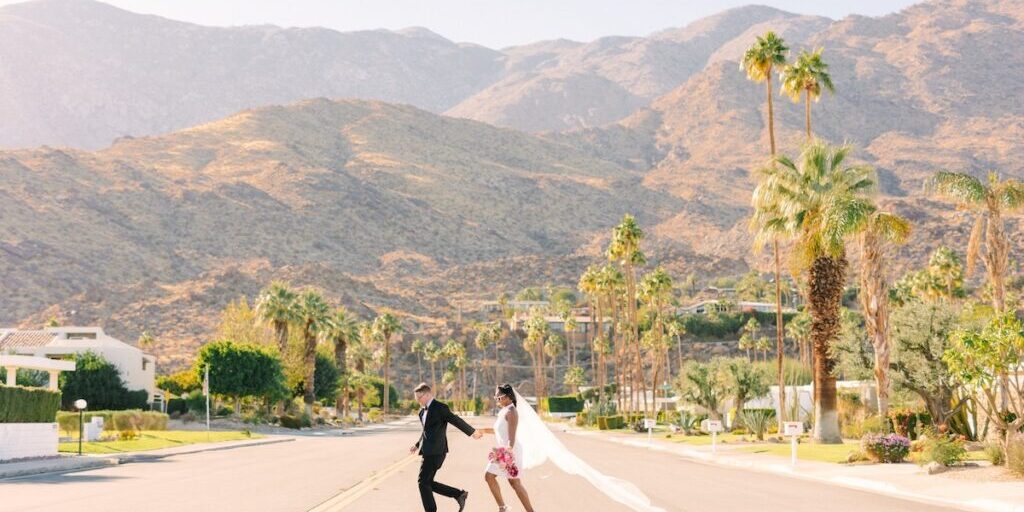A couple in wedding attire holds hands and runs down a palm-lined street with mountains in the background under a clear sky.