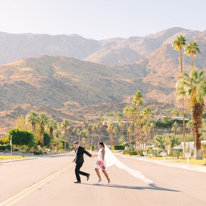 A couple in wedding attire holds hands and runs down a palm-lined street with mountains in the background under a clear sky.
