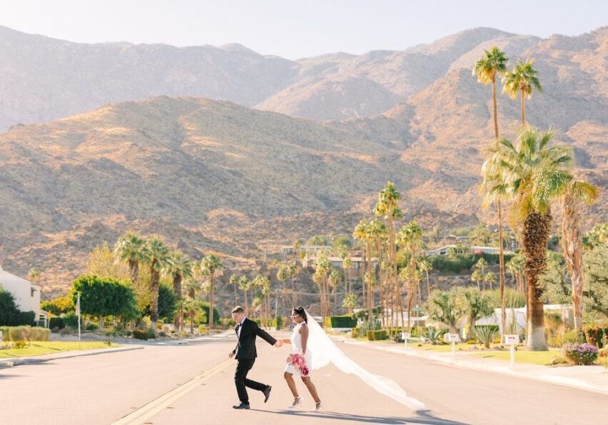 A couple in wedding attire holds hands and runs down a palm-lined street with mountains in the background under a clear sky.