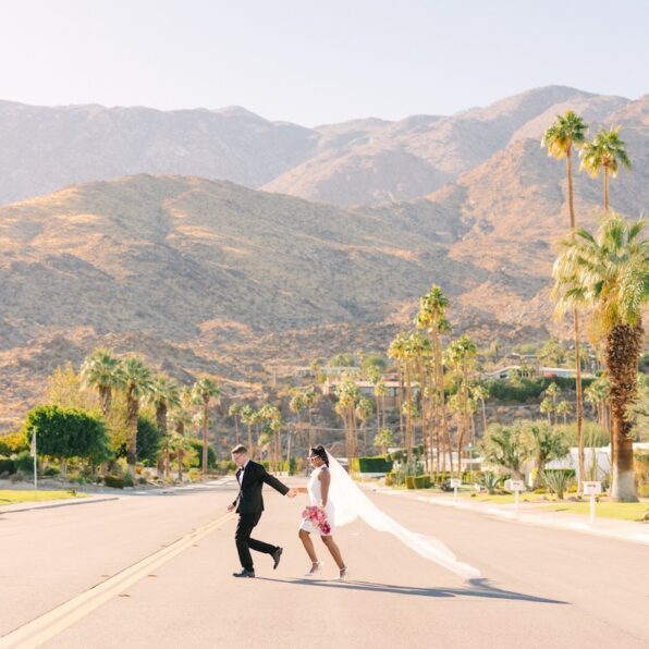 A couple in wedding attire holds hands and runs down a palm-lined street with mountains in the background under a clear sky.