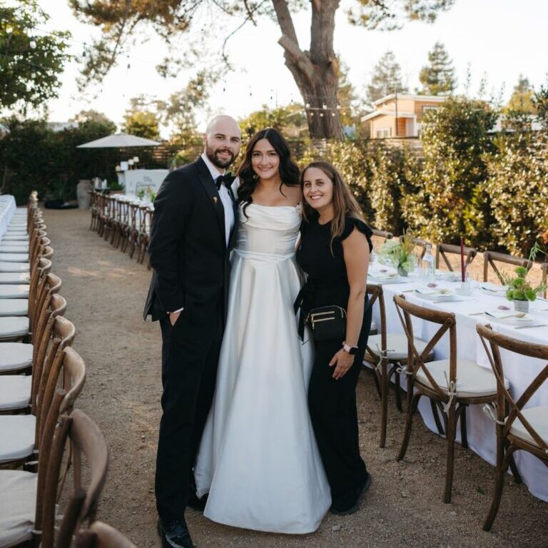 A bride in a white gown stands between two people, one in a suit and one in black, at an outdoor event with long tables and chairs.