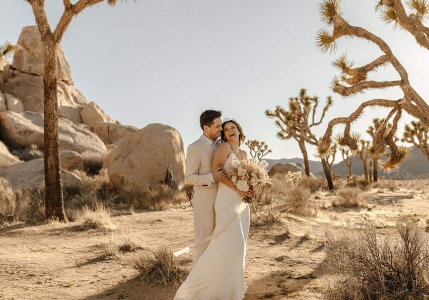 A couple dressed in wedding attire stands in a desert landscape with Joshua trees, sunlight casting shadows on the sandy ground.
