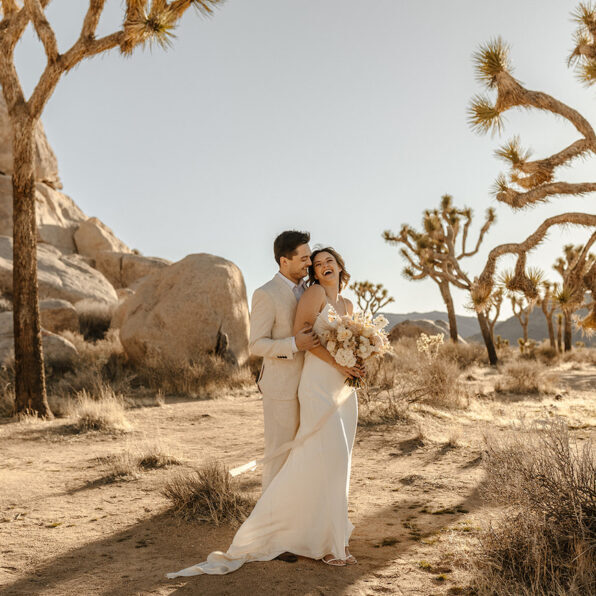 A couple dressed in wedding attire stands in a desert landscape with Joshua trees, sunlight casting shadows on the sandy ground.