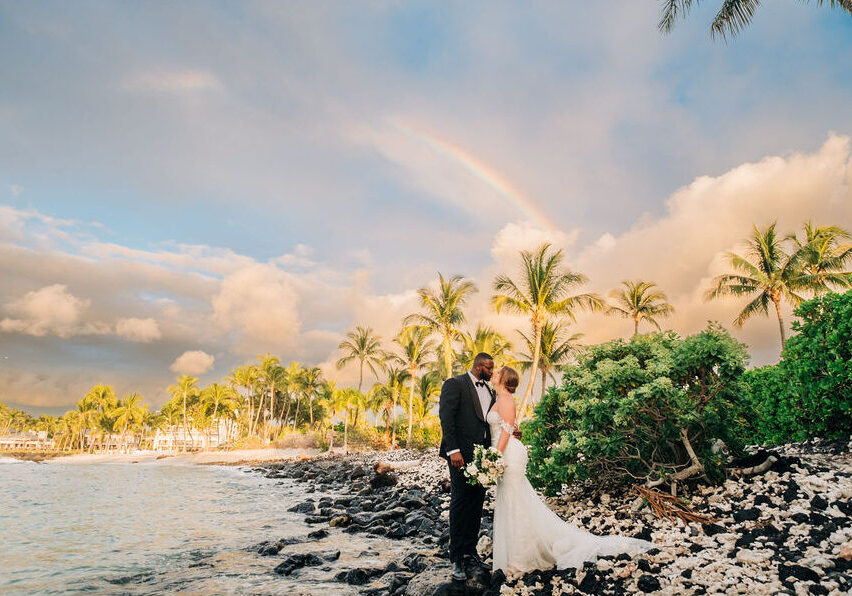 A bride and groom stand on a rocky beach with palm trees and a rainbow in the background. The bride holds a bouquet, and they are dressed in formal wedding attire.