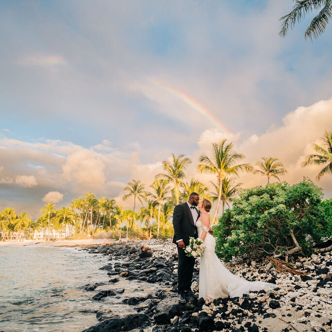 A bride and groom stand on a rocky beach with palm trees and a rainbow in the background. The bride holds a bouquet, and they are dressed in formal wedding attire.