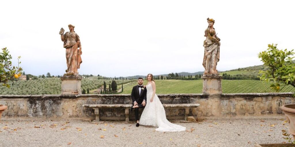 A bride and groom sit on a stone bench in an outdoor setting with statues and a scenic Tuscan landscape in the background.