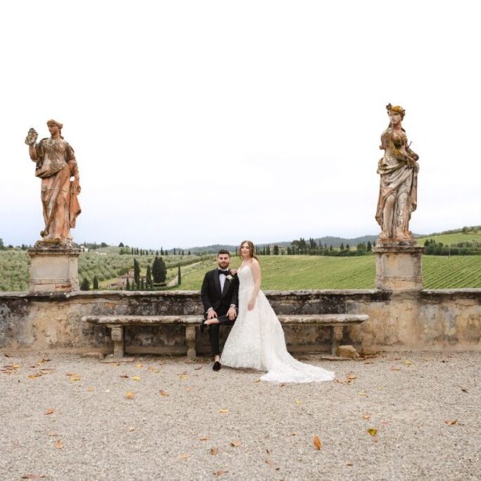 A bride and groom sit on a stone bench in an outdoor setting with statues and a scenic Tuscan landscape in the background.