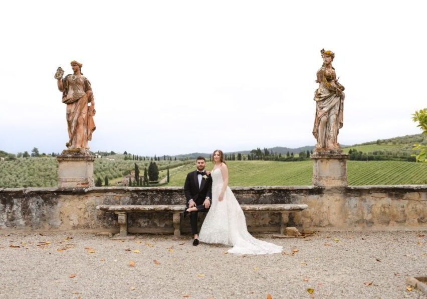 A bride and groom sit on a stone bench in an outdoor setting with statues and a scenic Tuscan landscape in the background.