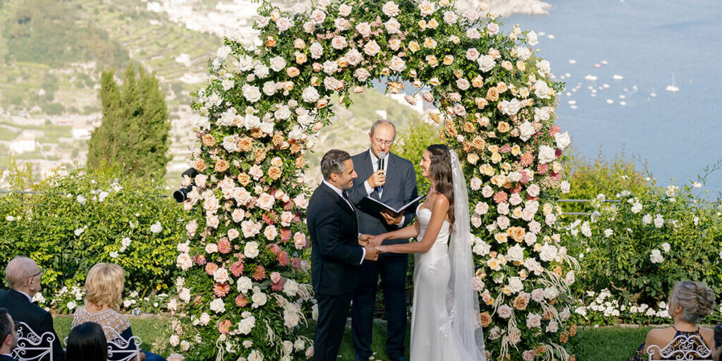 A couple stands under a large floral arch exchanging vows during an outdoor wedding ceremony overlooking a coastal landscape.