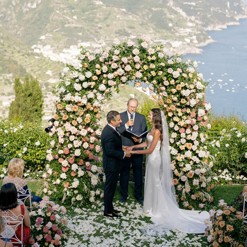 A couple stands under a large floral arch exchanging vows during an outdoor wedding ceremony overlooking a coastal landscape.