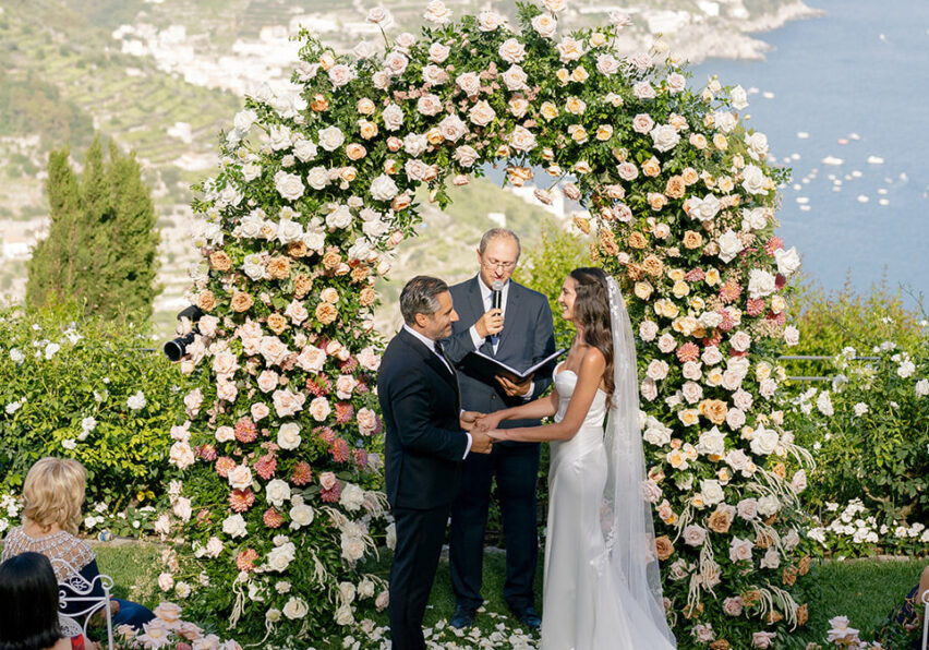 A couple stands under a large floral arch exchanging vows during an outdoor wedding ceremony overlooking a coastal landscape.