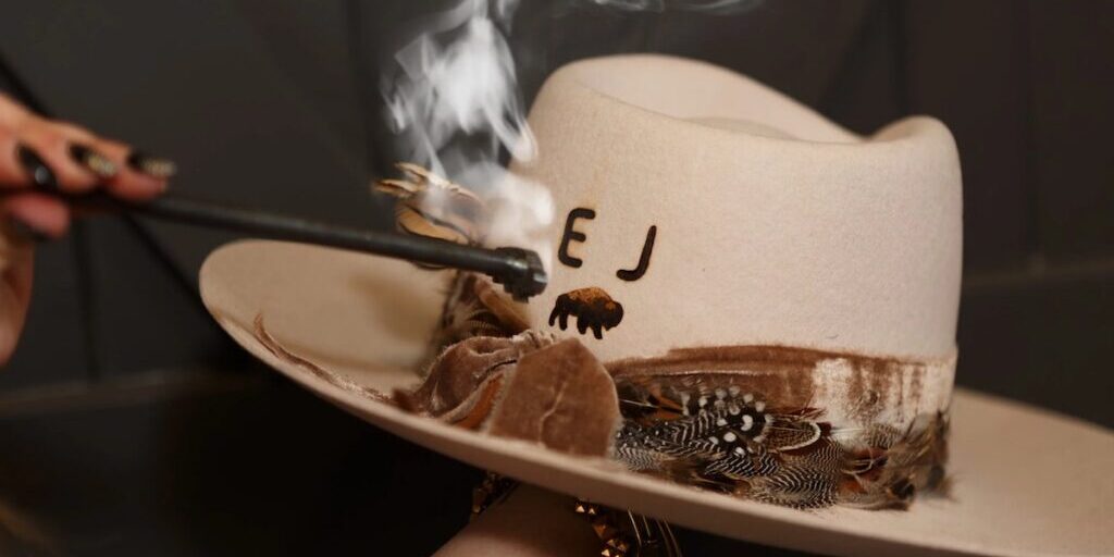A hand burns initials into a white cowboy hat with feathers and a decorative band, using a smoking branding tool.
