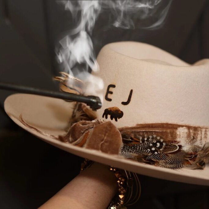 A hand burns initials into a white cowboy hat with feathers and a decorative band, using a smoking branding tool.