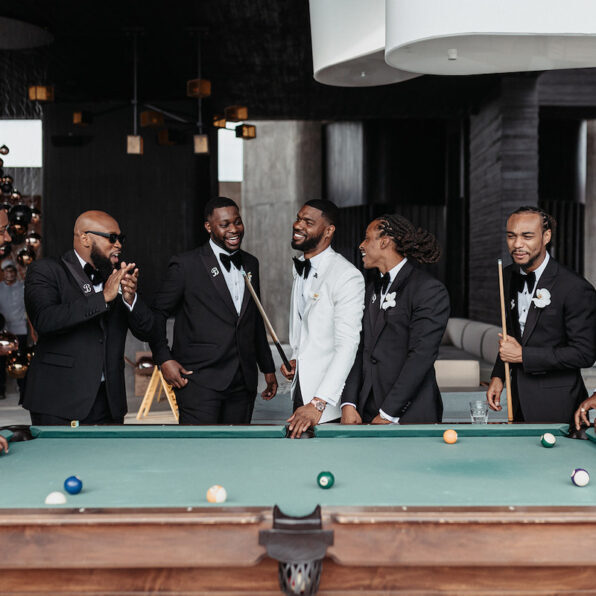 Five men in formal suits, one in white and four in black, stand around a pool table smiling and talking, holding pool cues in a modern indoor setting.