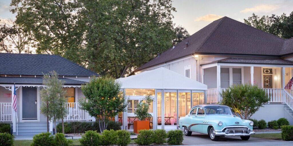 A vintage blue car is parked in front of a white house with a tented outdoor seating area; trees and shrubs line the sidewalk under a partly cloudy sky.