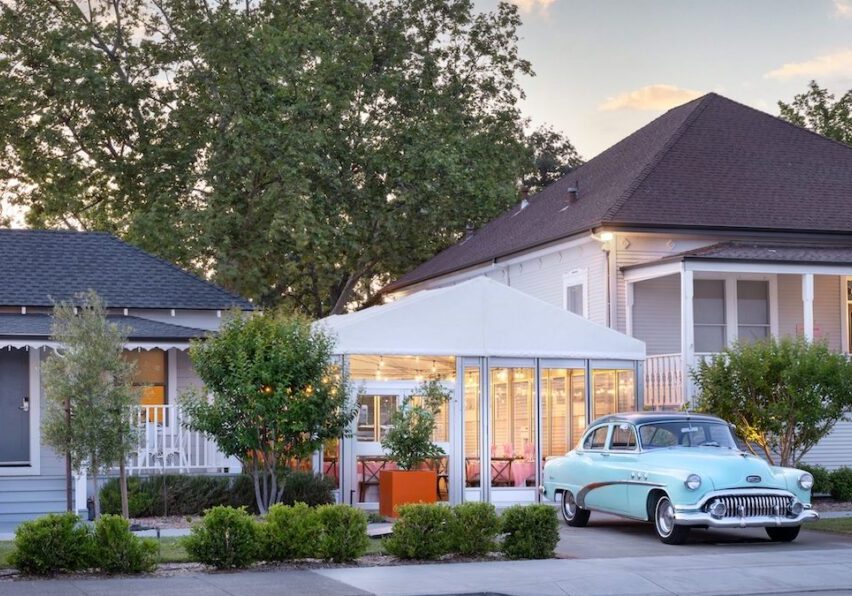A vintage blue car is parked in front of a white house with a tented outdoor seating area; trees and shrubs line the sidewalk under a partly cloudy sky.