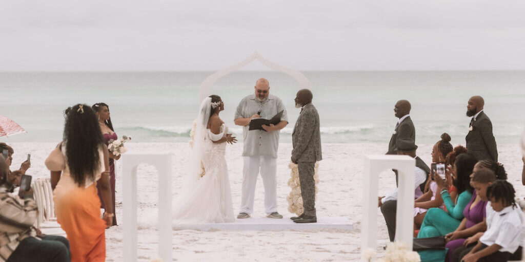 A bride and groom stand with an officiant under an arch on a sandy beach, surrounded by seated and standing guests, with the ocean in the background.