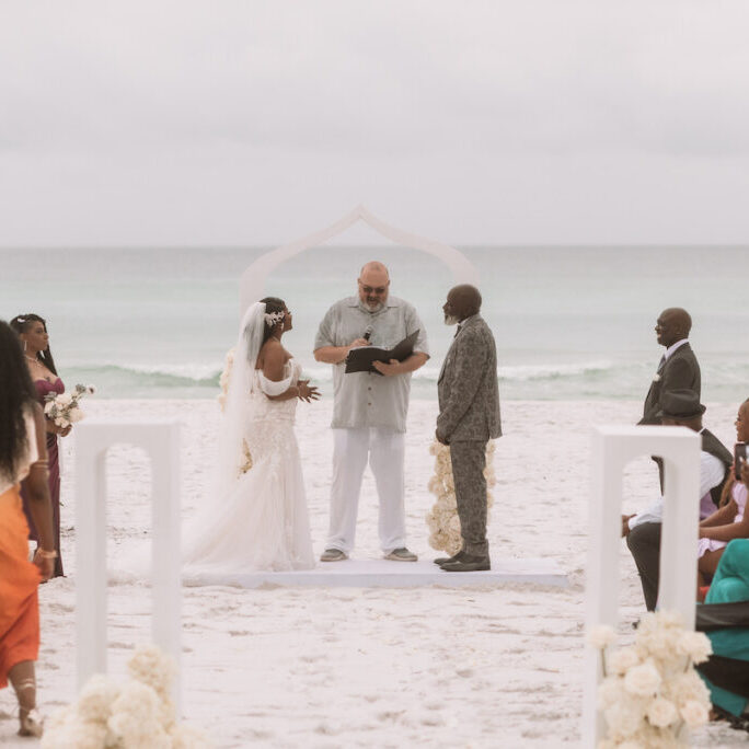 A bride and groom stand with an officiant under an arch on a sandy beach, surrounded by seated and standing guests, with the ocean in the background.