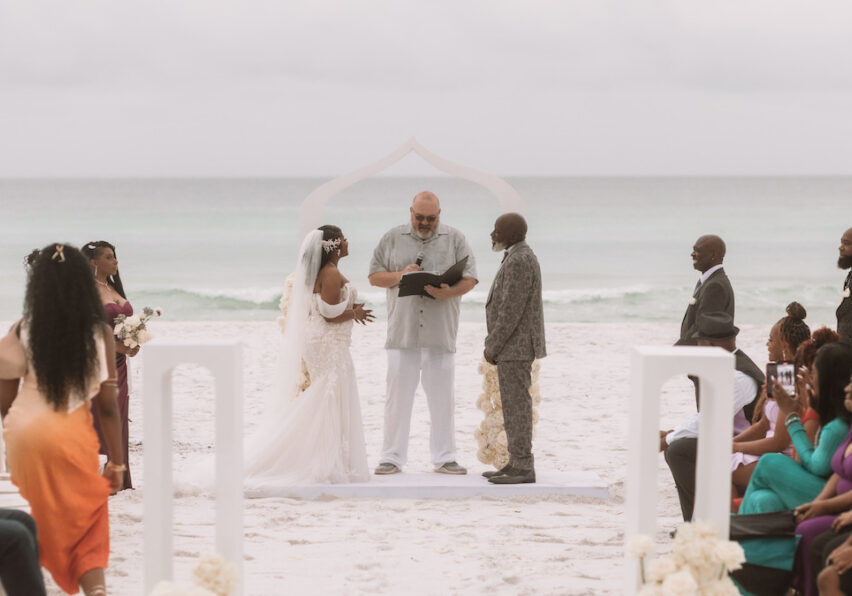A bride and groom stand with an officiant under an arch on a sandy beach, surrounded by seated and standing guests, with the ocean in the background.