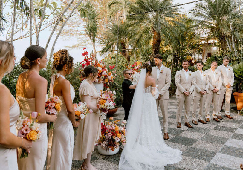 A bride and groom stand at the altar during an outdoor wedding ceremony, surrounded by bridesmaids and groomsmen in matching beige attire, with tropical plants in the background.