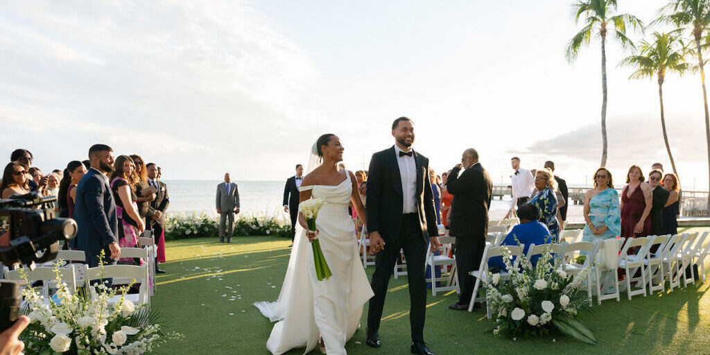 A bride and groom walk hand-in-hand down an outdoor aisle, smiling, with seated guests on either side and the ocean visible in the background.