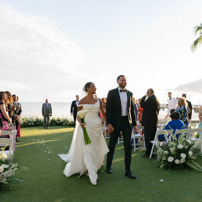 A bride and groom walk hand-in-hand down an outdoor aisle, smiling, with seated guests on either side and the ocean visible in the background.