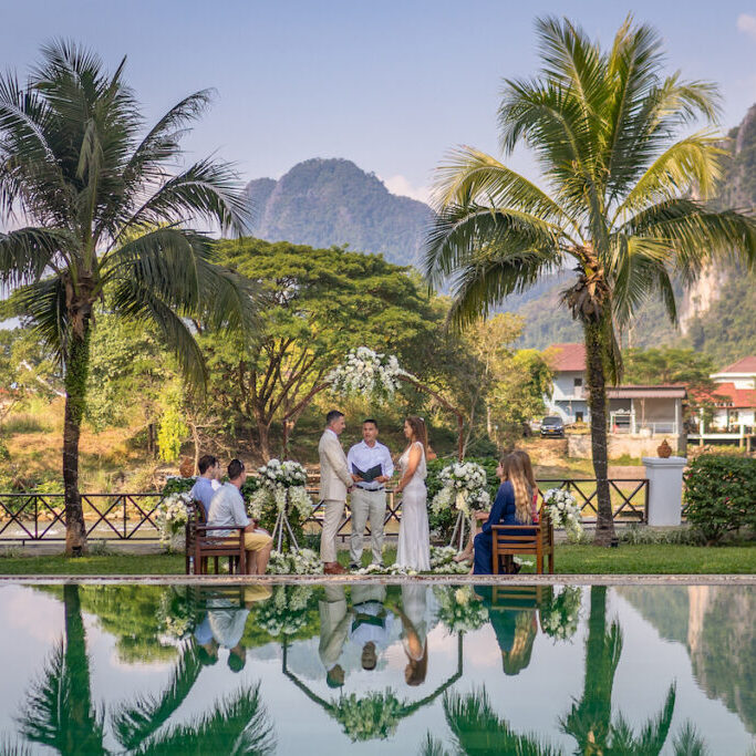 A couple is getting married outdoors near a pool, surrounded by palm trees, mountains, and a small group of people. The ceremony takes place in a tropical setting under a clear sky.