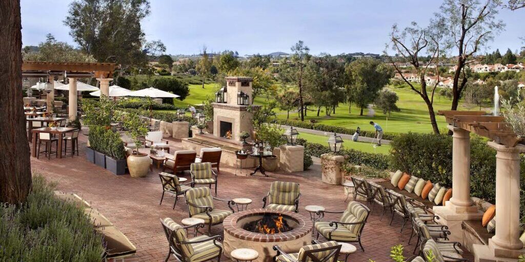 Outdoor patio with cushioned chairs around a fire pit, stone fireplace, and scenic view of a green lawn and trees in the background.