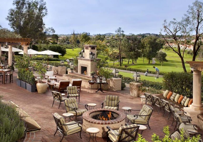 Outdoor patio with cushioned chairs around a fire pit, stone fireplace, and scenic view of a green lawn and trees in the background.