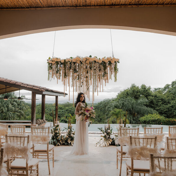 A bride in a white dress stands holding a bouquet under a floral chandelier in an outdoor wedding venue with empty decorated chairs and greenery in the background.