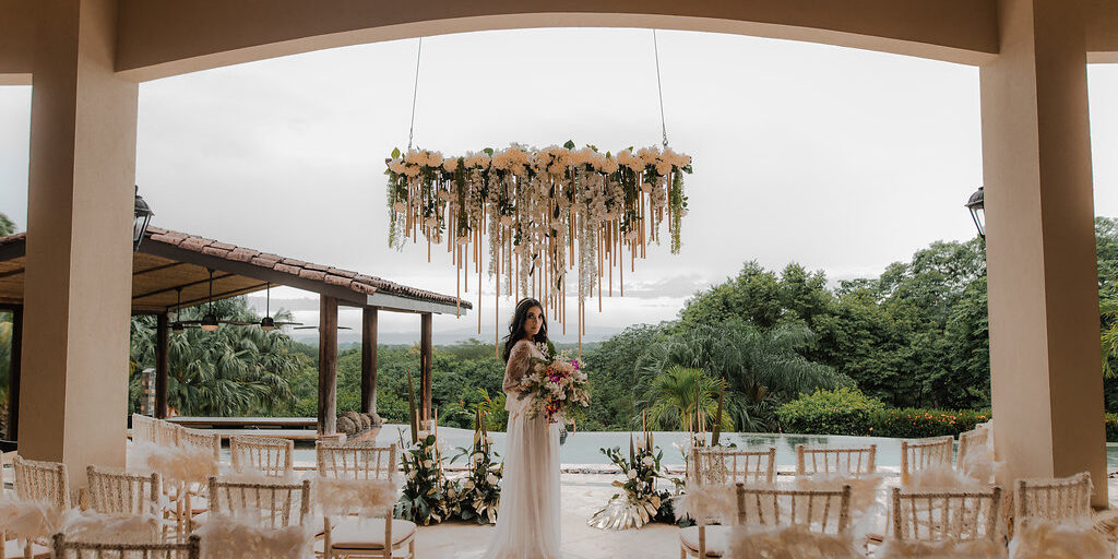A bride in a white dress stands holding a bouquet under a floral chandelier in an outdoor wedding venue with empty decorated chairs and greenery in the background.
