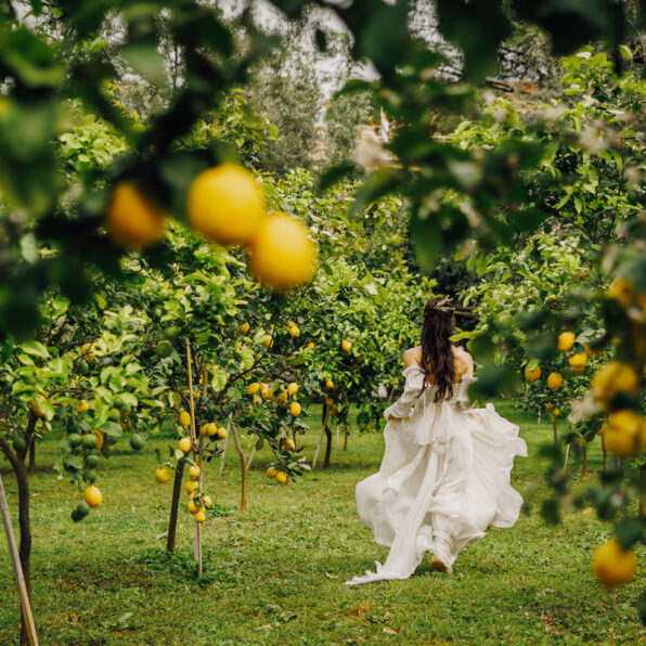 A person in a flowing white dress moves through a lemon orchard, surrounded by green foliage and yellow lemons.