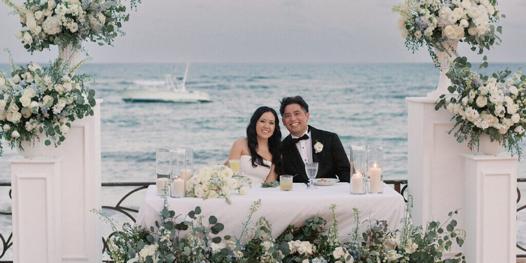 A bride and groom sit together at a flower-adorned table on a seaside terrace, smiling at the camera with the ocean and a boat in the background.