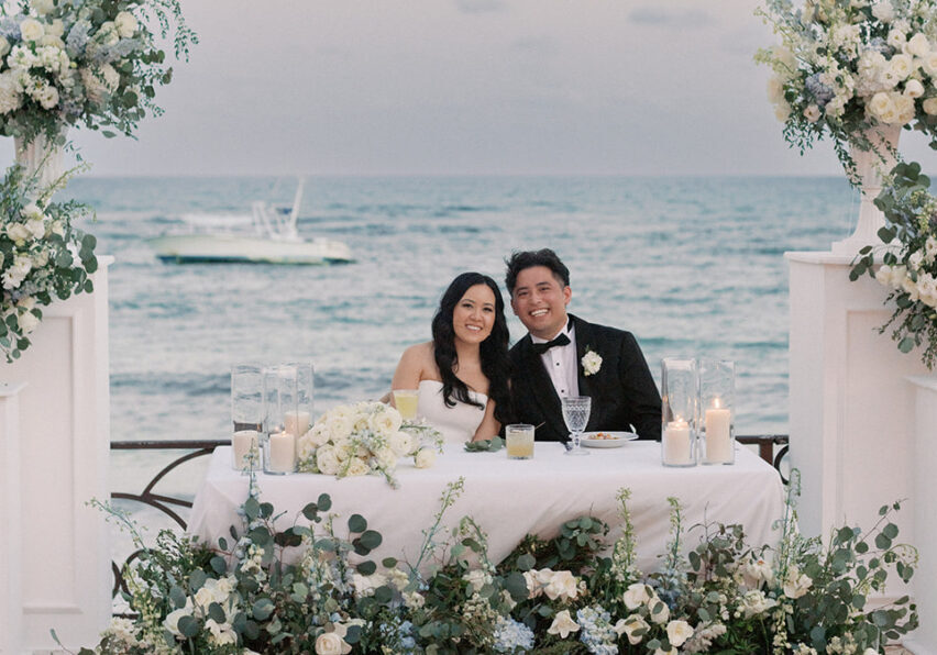 A bride and groom sit together at a flower-adorned table on a seaside terrace, smiling at the camera with the ocean and a boat in the background.