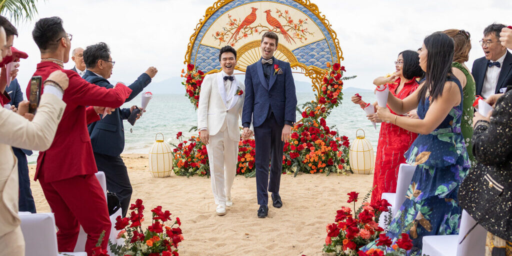 Two grooms walk hand in hand down an aisle on a beach, surrounded by guests and red floral decorations, with a decorative arch in the background.