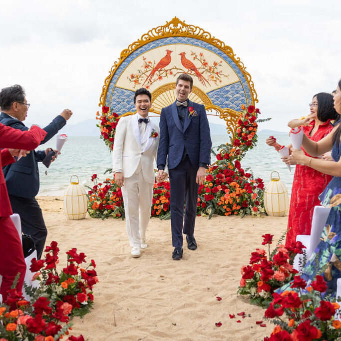 Two grooms walk hand in hand down an aisle on a beach, surrounded by guests and red floral decorations, with a decorative arch in the background.