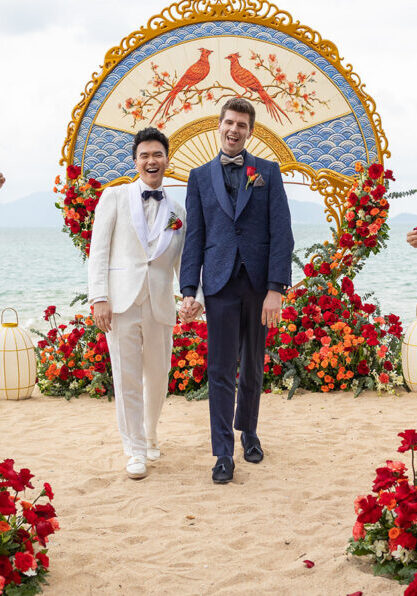 Two grooms walk hand in hand down an aisle on a beach, surrounded by guests and red floral decorations, with a decorative arch in the background.