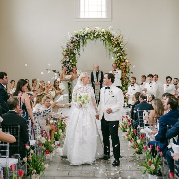 A bride and groom walk down the aisle, smiling, as guests throw flower petals in a decorated indoor wedding ceremony with floral arrangements and an arch.