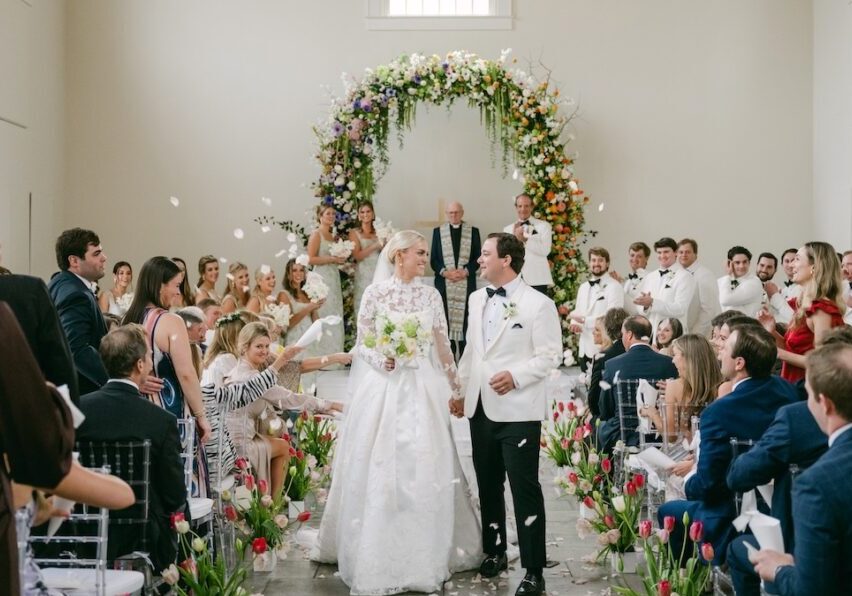 A bride and groom walk down the aisle, smiling, as guests throw flower petals in a decorated indoor wedding ceremony with floral arrangements and an arch.