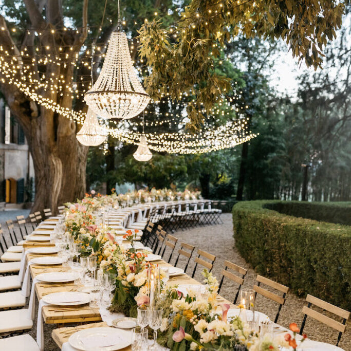 An outdoor long dining table is elegantly set with floral arrangements and tableware under hanging chandeliers and string lights.