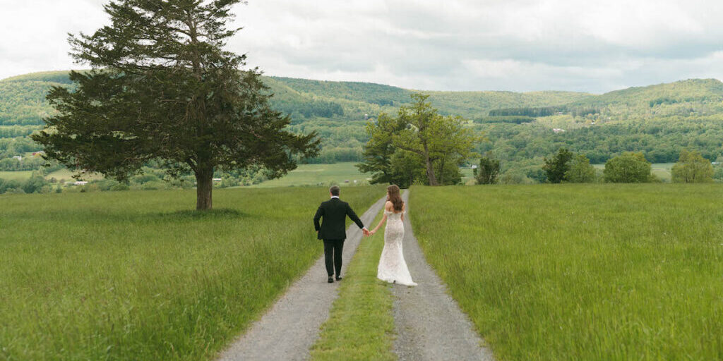 A bride and groom walk hand in hand down a gravel path through a green field, with hills and trees in the background under a cloudy sky.