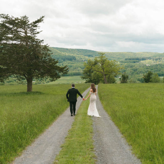 A bride and groom walk hand in hand down a gravel path through a green field, with hills and trees in the background under a cloudy sky.