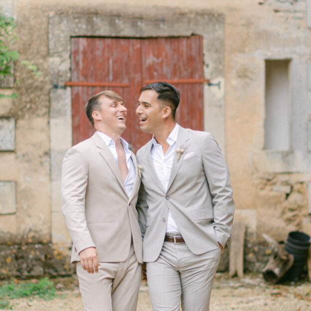 Noel and Daniel laugh together walking shoulder to shoulder away from an old French countryside building with a red door