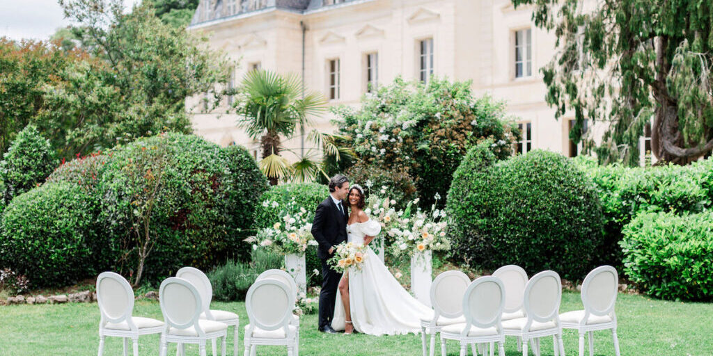 A bride and groom stand together in front of floral arrangements with empty chairs arranged on a lawn, outside a large, elegant building.