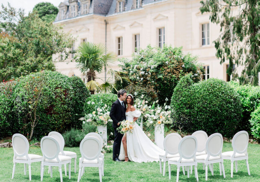 A bride and groom stand together in front of floral arrangements with empty chairs arranged on a lawn, outside a large, elegant building.