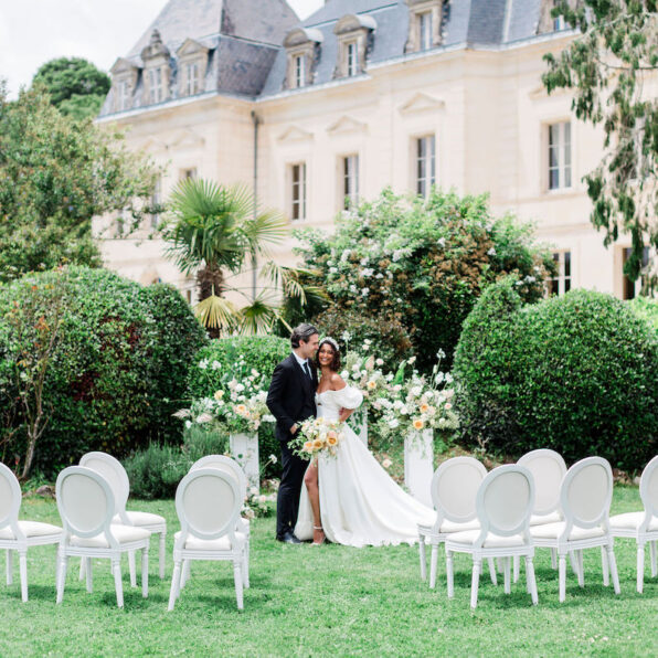 A bride and groom stand together in front of floral arrangements with empty chairs arranged on a lawn, outside a large, elegant building.