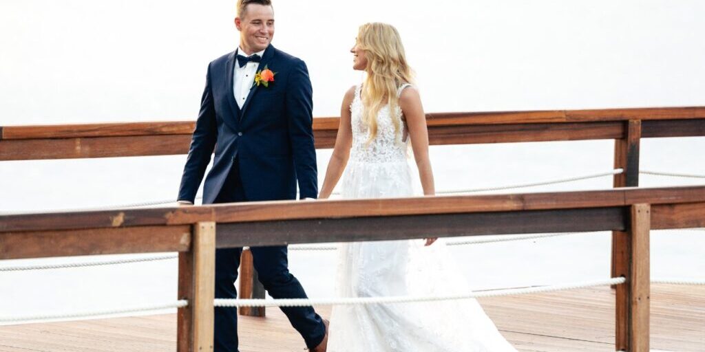 A couple in wedding attire walks hand in hand on a wooden deck.