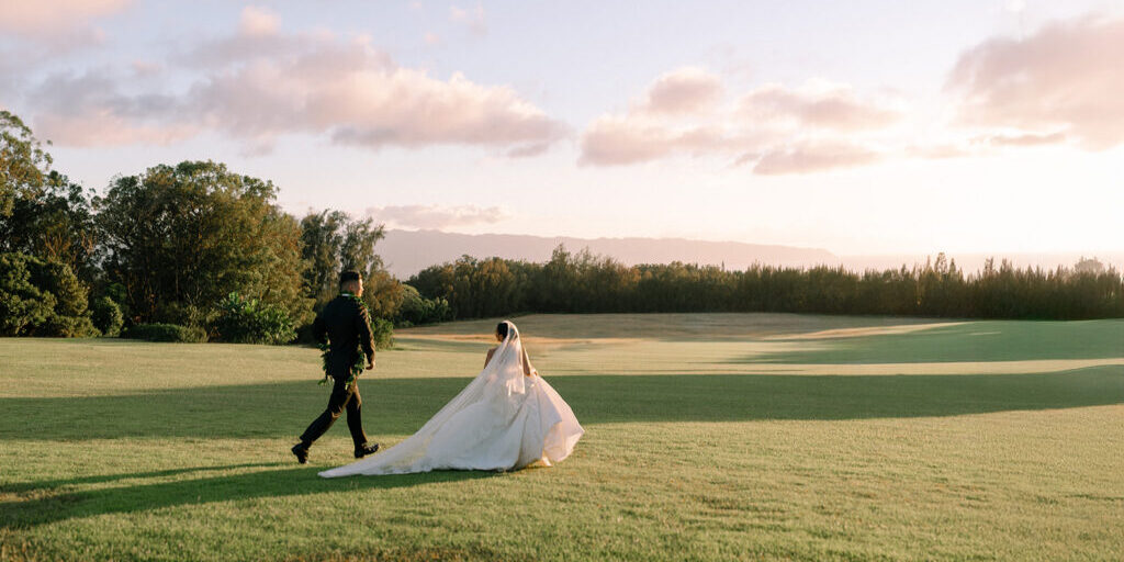 A bride and groom walk across a grassy field under a partly cloudy sky during sunset.