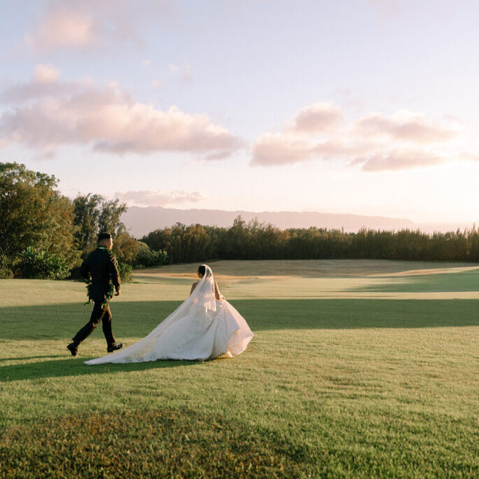 A bride and groom walk across a grassy field under a partly cloudy sky during sunset.